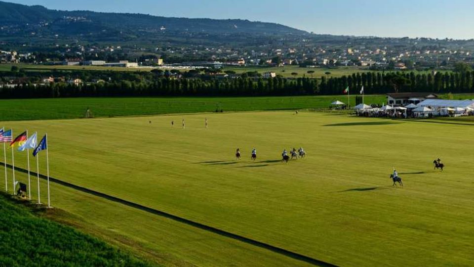 Ampio campo da polo in erba con bandiere su pennoni e strutture con tende e tribune a bordo campo. Sullo sfondo si vedono campi coltivati, una fila di alberi, un centro abitato e colline sotto un cielo sereno.