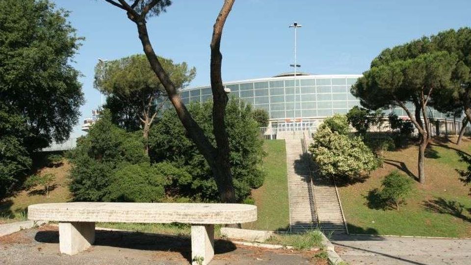 Vista di un parco su un pendio con alberi ad alto fusto e una panchina in cemento in primo piano. Una lunga scalinata centrale conduce a un edificio moderno con facciata curva in vetro sullo sfondo.