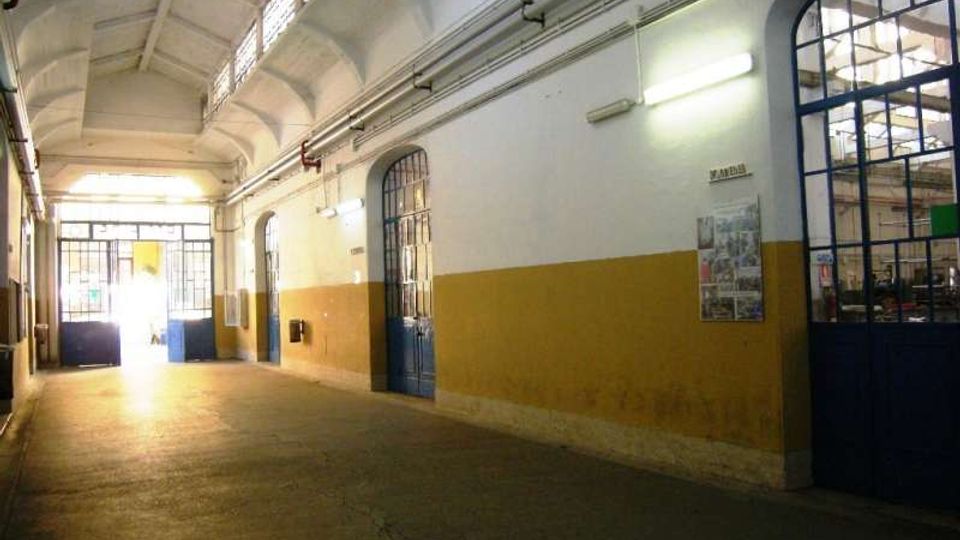 Corridoio interno di un edificio industriale con soffitto alto a volte, pareti bianche e fascia ocra, illuminazione al neon e tubazioni a vista lungo la parete. Grandi porte e finestre ad arco con telai metallici blu si aprono su altri ambienti e verso un ingresso luminoso in fondo.