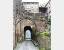 Strada acciottolata in un borgo storico che attraversa un grande arco in pietra, formando un passaggio coperto tra edifici in muratura. Muri con muschio e rami rampicanti circondano il tunnel, con una vista sulla valle visibile in lontananza oltre l’uscita.