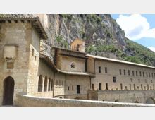 Complesso monastico in pietra con arcate, finestre regolari e piccolo campanile, addossato a una parete rocciosa. In primo piano un muretto e sullo sfondo una montagna boscosa sotto cielo parzialmente nuvoloso.
