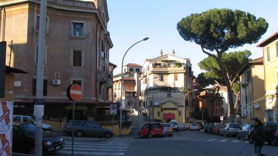 Incrocio urbano con strisce pedonali e auto parcheggiate lungo la strada, circondato da edifici residenziali di più piani con facciate in intonaco. Sullo sfondo si vede una scalinata che sale tra le palazzine e un grande pino mediterraneo vicino al margine destro.