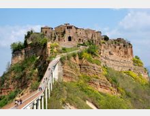 Borgo storico in pietra arroccato sulla sommità di una rupe di tufo, con case e una chiesa al centro. Un lungo ponte pedonale sopraelevato conduce all’ingresso, circondato da pendii verdi e stratificazioni rocciose sotto un cielo parzialmente nuvoloso.