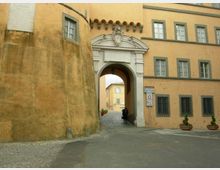 Ingresso ad arco in pietra con frontone decorato e stemma, inserito in un edificio intonacato color ocra con finestre rettangolari. Un passaggio conduce a un cortile interno con pavimentazione in ciottoli, affiancato da una torre cilindrica e da vasi con piante.