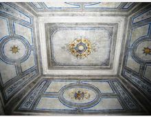 Soffitto interno decorato con riquadri geometrici bordati in blu e cornici in stucco, con pannello centrale rettangolare e medaglioni ornamentali dorati. La superficie è dipinta in tonalità chiare con motivi floreali e rosoni simmetrici.