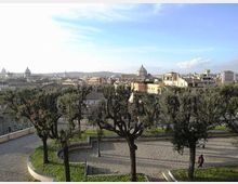 Veduta panoramica su una città con numerose cupole e campanili all’orizzonte, sotto un cielo limpido. In primo piano si vede un belvedere con alberi potati, vialetti curvi in pietra e una zona verde.