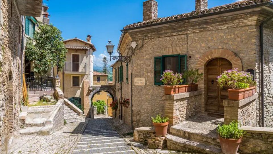 Stradina in pietra lastricata in un borgo storico, con scalinate e case in muratura dai tetti in coppi e persiane verdi. In primo piano si vedono un portale ad arco, un lampione e terrazze con vasi di fiori e piante lungo i muri.