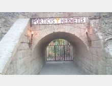 Ingresso a tunnel ad arco in pietra con volta in mattoni e pavimento in cemento, sormontato dall’insegna “PORTICVS HERMETIS”. Ai lati dell’arco sono fissate due torce e in fondo si vede un cancello in ferro che chiude il passaggio verso un’area alberata.