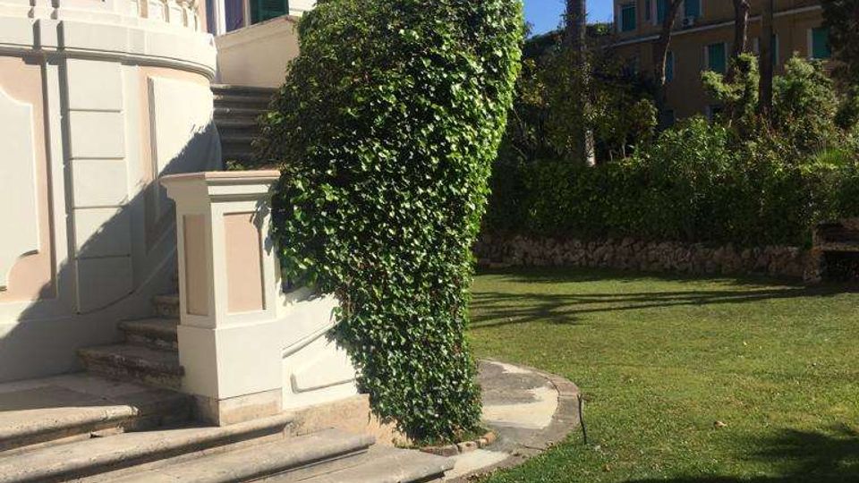 Esterno di una villa in stile classico con facciata chiara, persiane verdi e scalinata curva con balaustra. In primo piano un prato curato con siepe potata e, sullo sfondo, palme e altri edifici tra la vegetazione sotto un cielo sereno.