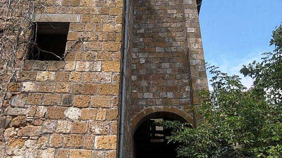 Facciata laterale di un edificio storico in pietra con finestre rettangolari e un passaggio ad arco che conduce a un corridoio ombreggiato. Vegetazione fitta sul lato destro e cielo azzurro sullo sfondo.