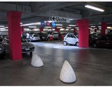 Parcheggio sotterraneo multipiano con colonne strutturali rosse, file di auto in sosta e illuminazione a neon sul soffitto in cemento. Al centro sono visibili cartelli stradali sospesi con la scritta “Uscita” e indicazioni di direzione.