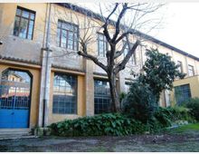 Facciata di un edificio a due piani con intonaco giallo e grigio, grandi finestre rettangolari e portone blu con vetri. Davanti si trova un cortile con un grande albero spoglio e aiuole con arbusti e vegetazione bassa.