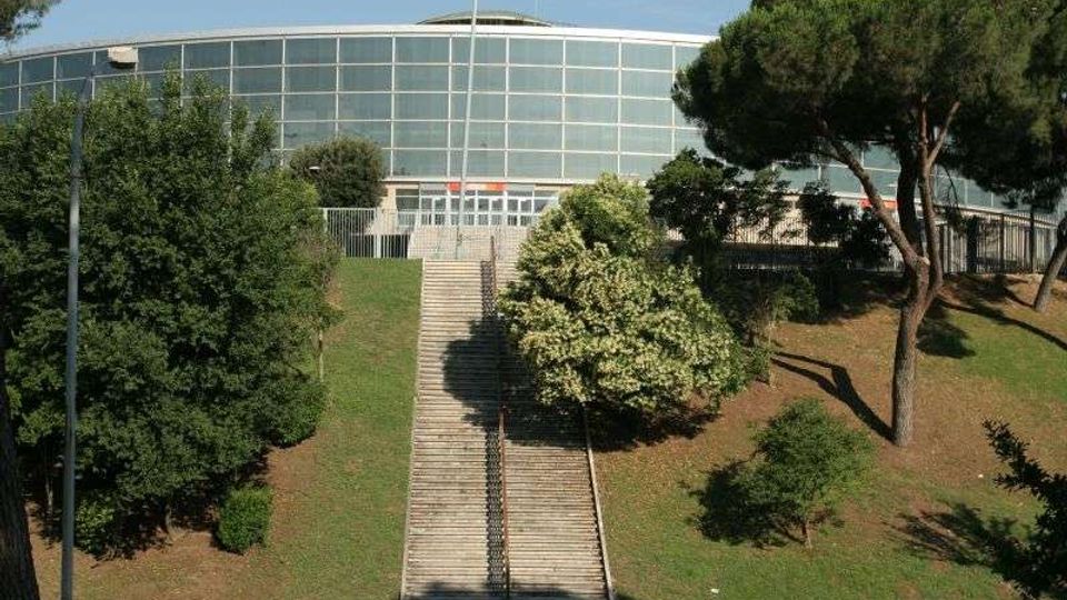 Edificio moderno a pianta semicircolare con ampia facciata vetrata, situato in cima a una scalinata centrale che risale un pendio erboso. Ai lati della scalinata ci sono alberi e cespugli, con lampioni e recinzioni lungo l’ingresso.