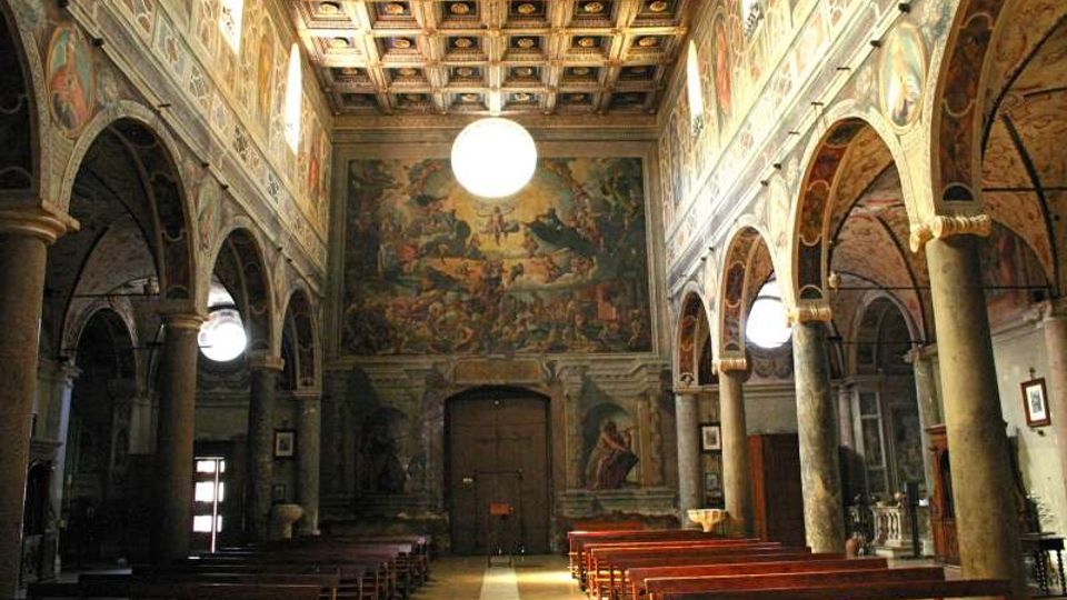 Interno di una chiesa con navata centrale fiancheggiata da arcate su colonne in pietra e file di panche in legno. Le pareti e la controfacciata sono riccamente affrescate, con soffitto a cassettoni decorato e grandi lampade sferiche sospese.