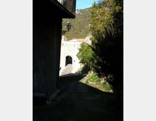 Vicolo in pendenza accanto a un edificio in pietra, con una lanterna a muro, che conduce a un arco in muratura illuminato dal sole. Sullo sfondo si vedono alberi e montagne boscose.