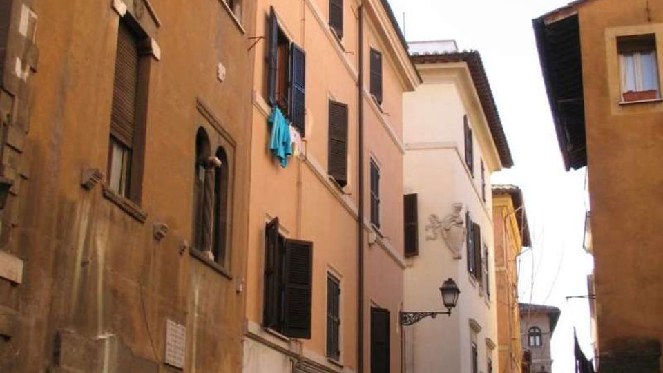 Strada stretta in un centro storico, fiancheggiata da alti edifici in intonaco e mattoni con persiane scure e finestre ravvicinate. Al piano terra si vedono archi e serrande chiuse, lampioni a parete e alcuni panni stesi tra le finestre.
