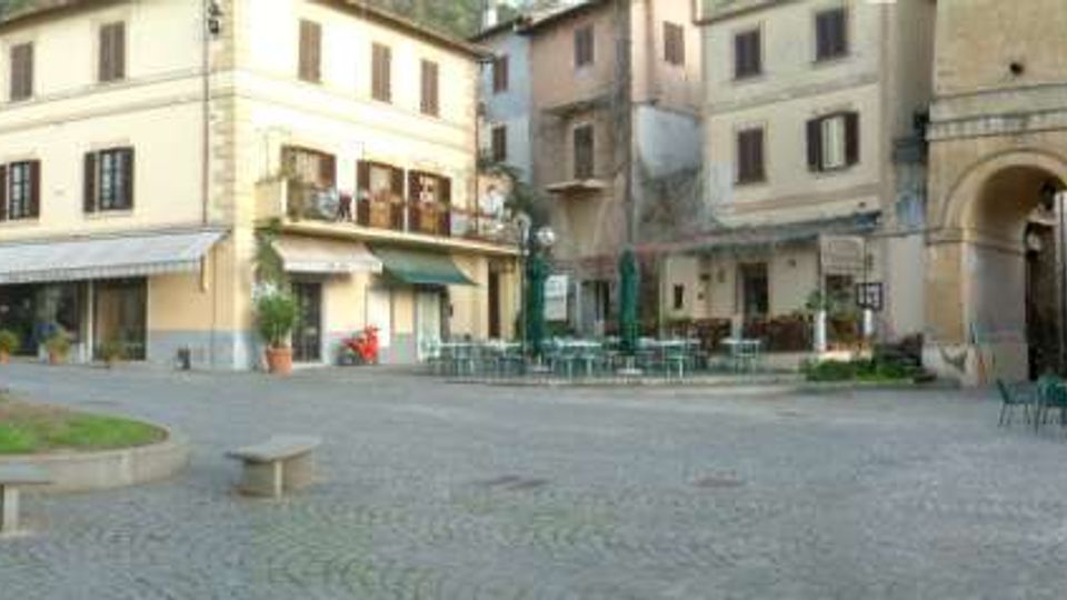 Piazza lastricata in un borgo storico, circondata da edifici in muratura con persiane e balconi. Sono visibili un portico ad arco, tavolini e sedie di un bar con ombrelloni chiusi e una grande palma con panchine in pietra.
