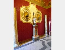 Interno di un edificio storico con pareti rosse damascate e pavimento in marmo a motivi geometrici. Sulla parete è fissata una grande targa decorativa dorata con nicchie a conchiglia e scritte, affiancata da due colonne in marmo e da un piedistallo con scultura.