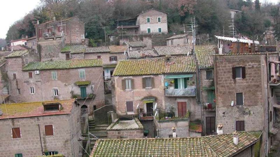 Vista panoramica di un borgo collinare con case in pietra addossate tra loro e tetti in coppi. Sullo sfondo si vede un pendio alberato con alcune abitazioni sparse e antenne sui tetti.