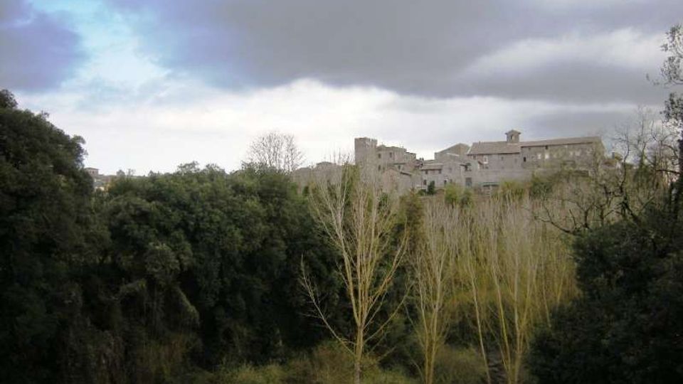 Veduta di un borgo in pietra su un’altura, con edifici compatti e una torre visibili sullo sfondo. In primo piano una valle con fitta vegetazione e alberi spogli, sotto un cielo nuvoloso.