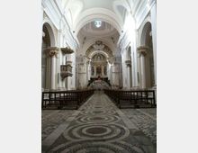 Interno di una chiesa con navata centrale lunga, pavimento a mosaico con motivi circolari e file di panche in legno ai lati. Alti archi e colonne bianche sorreggono volte decorate, con altare rialzato e cupola ornata sullo sfondo e una finestra circolare in alto.