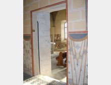 Passaggio ad arco che conduce a un interno di chiesa o cappella, con pareti affrescate e decorazioni geometriche. Si intravedono una finestra ad arco, un banco in legno e pavimento in pietra, con cornici murarie dipinte attorno all’apertura.