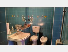 Piccolo bagno con pareti rivestite di piastrelle blu, lavabo bianco con specchio e dispenser di sapone. Sono presenti WC con cassetta esterna e bidet, con adesivi decorativi e una mensola con riviste sullo sfondo.