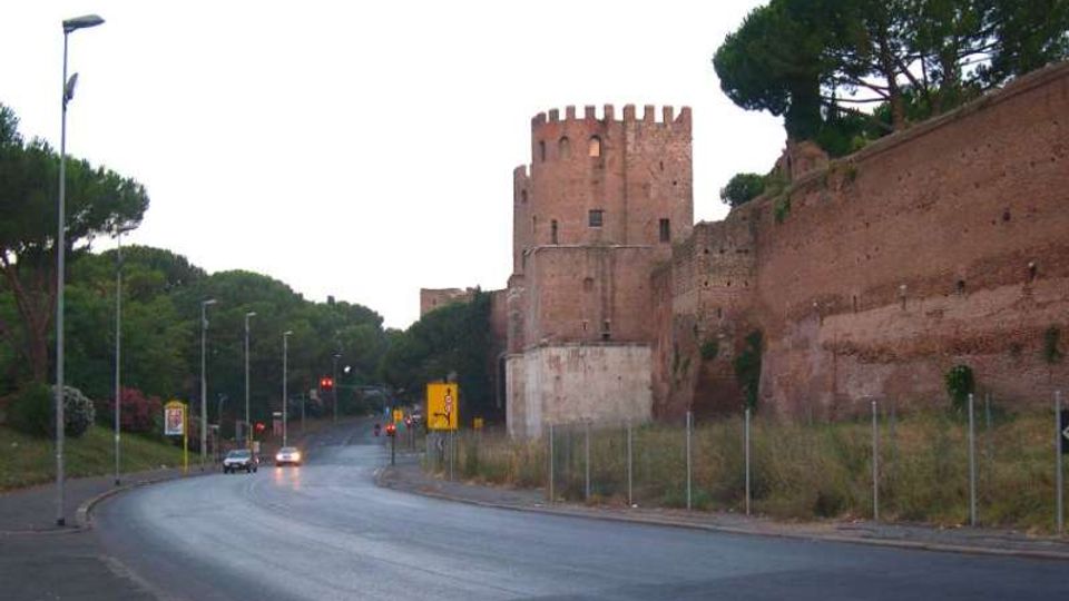 Strada asfaltata in curva con lampioni e segnaletica, affiancata da alte mura in mattoni e una torre cilindrica merlata su un basamento chiaro. Sullo sfondo si vedono alberi ad alto fusto e una zona erbosa recintata lungo il bordo della carreggiata.
