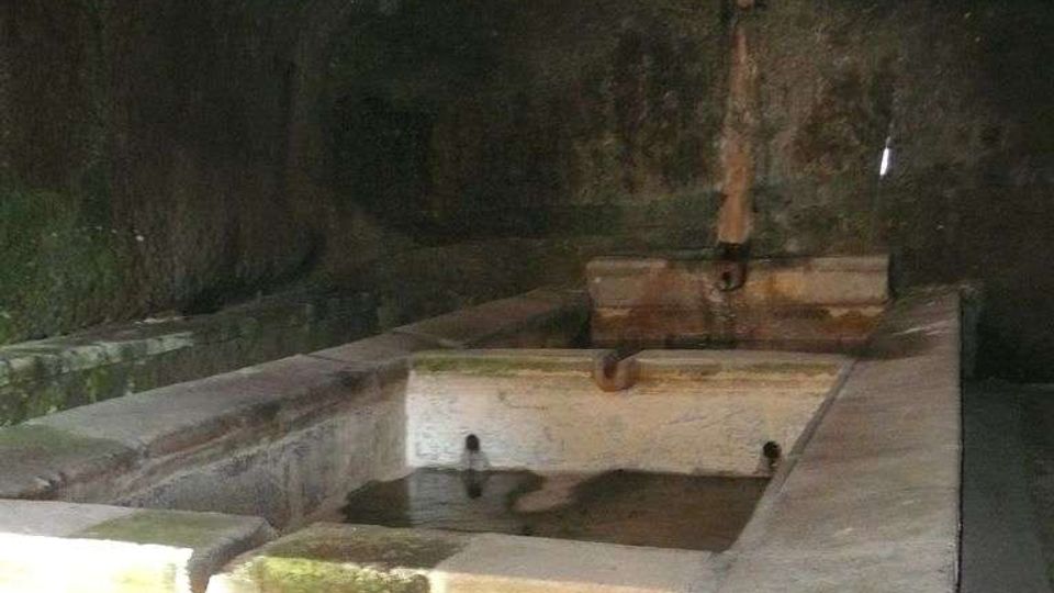 Interno in penombra di un antico lavatoio in muratura, con vasche rettangolari in pietra e acqua stagnante. La struttura è coperta da una volta bassa e presenta pareti consumate dall’umidità e un rubinetto centrale.