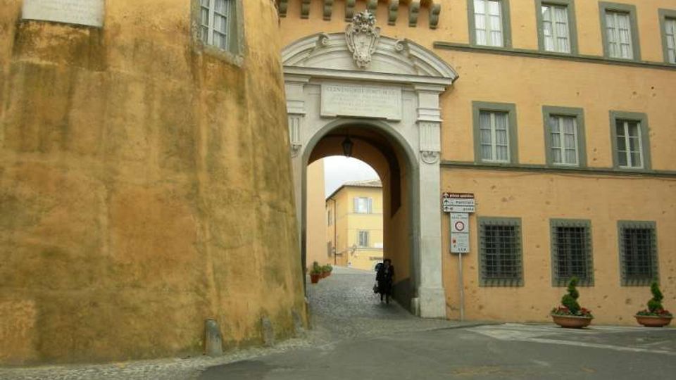 Ingresso ad arco in pietra con frontone decorato e stemma, inserito in un edificio intonacato color ocra con finestre rettangolari. Un passaggio conduce a un cortile interno con pavimentazione in ciottoli, affiancato da una torre cilindrica e da vasi con piante.