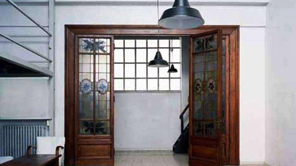 Interno di un edificio con ampio spazio vuoto e pavimento in piastrelle chiare, illuminato da lampade industriali a sospensione. Al centro c’è un grande varco con porte in legno e vetrate decorate, che conduce a un’area con finestra a griglia e una scala laterale; a sinistra si vede una struttura metallica a più livelli.