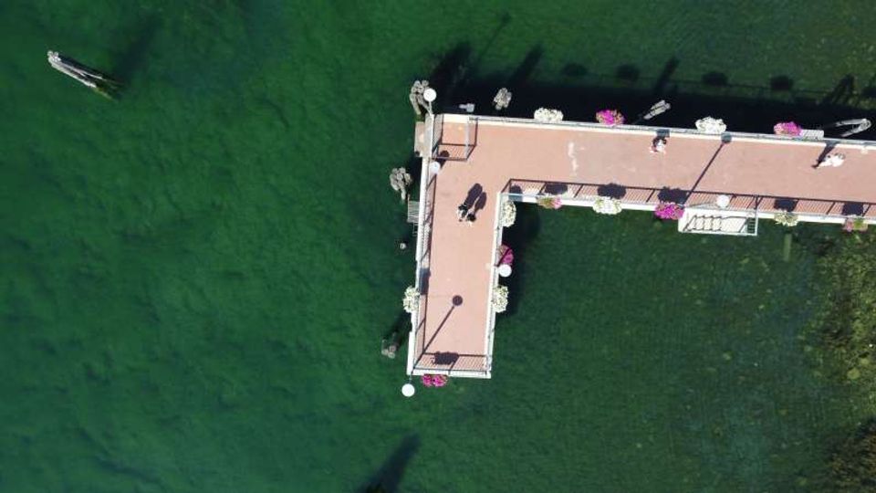 Veduta dall’alto di un molo a forma di L con pavimentazione rosa e ringhiere, decorato con fioriere, che si estende su acqua verde scura. Si notano alcune piccole imbarcazioni e le ombre delle strutture riflesse sull’acqua.