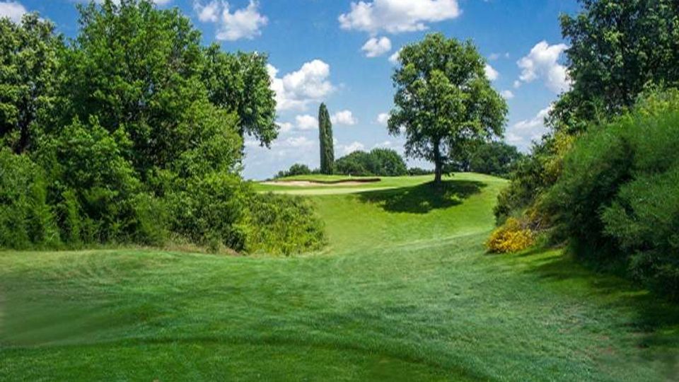 Ampio campo da golf con prato curato e lievi ondulazioni, circondato da alberi e cespugli. Sullo sfondo si vedono un green con bunker di sabbia e un alto cipresso sotto un cielo azzurro con nuvole sparse.