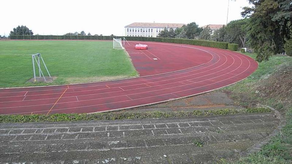 Pista di atletica in gomma rossa che curva attorno a un campo in erba verde, con linee di corsia e alcune attrezzature sportive. Sullo sfondo si vedono alberi e un grande edificio chiaro, con una scarpata e una gradinata in cemento in primo piano.