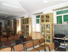 Aula scolastica con file di sedie rivolte verso una lavagna su carrello, pavimento chiaro e illuminazione a neon sul soffitto. Lungo le pareti ci sono alte librerie con ante in vetro e finestre con tapparelle verdi.