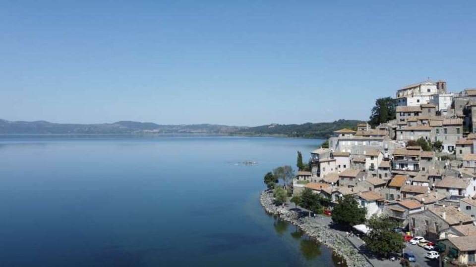 Veduta panoramica di un grande lago calmo con colline basse all’orizzonte. Sulla riva a destra si trova un borgo compatto di case in pietra con tetti in coppi, disposto in salita fino a un edificio più grande in cima, con una strada e alberi lungo il lungolago.