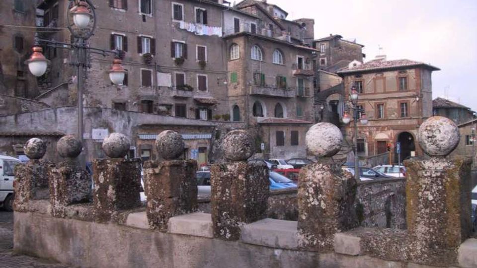 Balaustra in pietra con pilastrini sormontati da sfere in primo piano, lungo una piazza lastricata. Sullo sfondo si vedono edifici storici addossati su un pendio, con facciate in muratura, finestre e tetti in tegole, oltre a lampioni e auto parcheggiate.