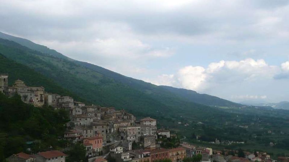 Veduta panoramica di un borgo collinare con case in pietra e tetti in tegole addossate al pendio, ai piedi di una catena montuosa. Sullo sfondo si estende una valle verde con campi e boschi sotto un cielo nuvoloso.