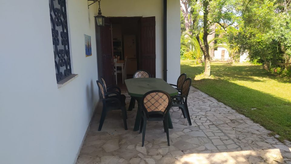 Portico di una casa con pareti bianche e tettoia, pavimentato in pietra, con tavolo rettangolare e sedie da esterno vicino a una porta in legno aperta. A destra si vede un giardino erboso con alberi e vegetazione, illuminato dalla luce del sole.