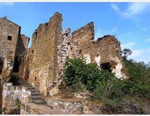 Rovine di un edificio in pietra con muri parzialmente crollati e intonaco consumato, affiancate da una scalinata in pietra. Vegetazione fitta cresce alla base e all’interno delle strutture, con cielo azzurro sullo sfondo.