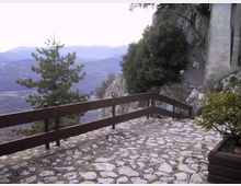 Terrazza panoramica con pavimentazione in pietra irregolare e parapetto in legno, affacciata su una valle montuosa. A lato si vedono parete rocciosa, alberi e un edificio in muratura addossato alla scogliera.