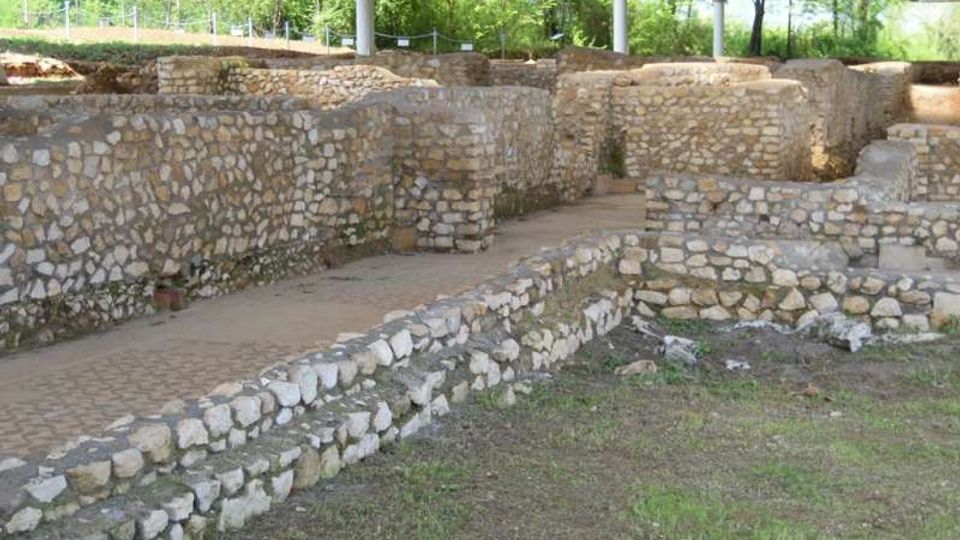 Scavi archeologici all’aperto con muri in pietra a bassa altezza e percorsi tra i resti, coperti da una tettoia metallica sostenuta da pilastri. Vegetazione e alberi circondano l’area sullo sfondo.