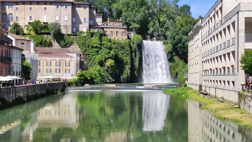 Veduta urbana lungo un fiume calmo con una grande cascata che scende da una scarpata verde. Ai lati si vedono edifici storici su una collina e un lungo edificio moderno sul lungofiume, con riflessi nell’acqua.