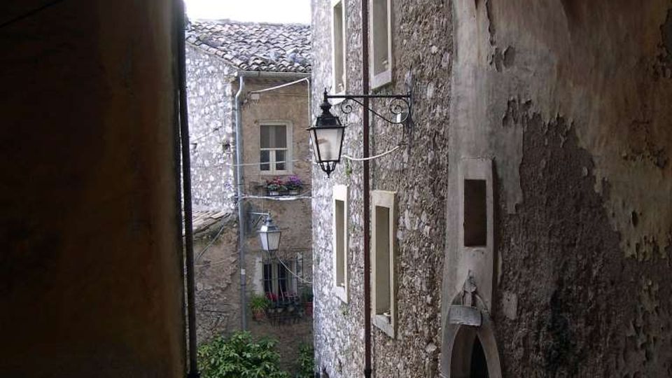 Passaggio ad arco in pietra che si apre su una stretta viuzza in discesa lastricata, tra edifici in pietra con finestre piccole e portale ad arco. Lanterne in ferro battuto sporgono dalle pareti e si intravedono tetti in coppi e alcune piante in fondo al vicolo.