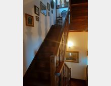 Scala interna in legno con corrimano e balaustra intagliata che sale a un pianerottolo superiore. Pareti bianche decorate con quadri e piatti, illuminate da una lampada a parete.