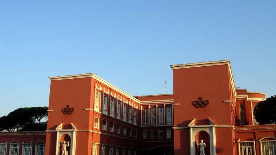 Edificio istituzionale in stile razionalista con facciata rosso mattone, due corpi laterali simmetrici e finestre verticali su più piani. All’ingresso ci sono nicchie con statue e un’ampia area frontale aperta sotto un cielo sereno, con alberi sullo sfondo.