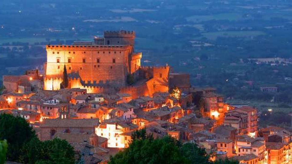 Veduta panoramica al crepuscolo di un borgo collinare con un grande castello medievale fortificato illuminato in cima, circondato da case in pietra dai tetti bassi. Sullo sfondo si estende una pianura con campi e strade, con vegetazione in primo piano.