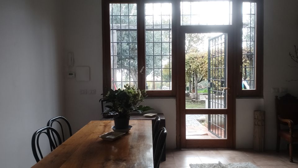Interno di una sala da pranzo con tavolo in legno e sedie scure, accanto a grandi finestre e a una porta vetrata con inferriata che si apre su un patio e un giardino. Pareti bianche, pavimento piastrellato e tappeto chiaro vicino all’ingresso.