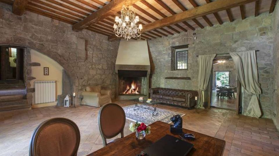 Ampio soggiorno rustico con pareti in pietra, soffitto con travi in legno a vista e lampadario in cristallo. Al centro c’è un grande camino acceso, con divani e tavolino su tappeto, e un passaggio ad arco con tende che conduce a un’altra stanza.