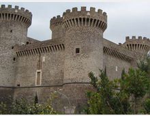 Castello medievale in pietra con mura fortificate e tre grandi torri cilindriche merlate, dotate di beccatelli e piccole feritoie. In primo piano si vede vegetazione arbustiva, con cielo coperto sullo sfondo.